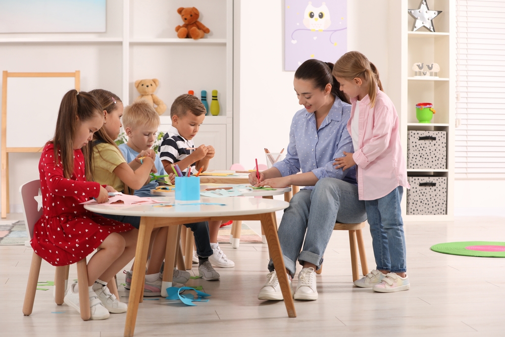 NurseryTeacherAndGroupOfCuteLittleChildrenMakingToys Teacher guiding a craft activity as children sit around a round table, coloring and cutting paper in a bright classroom with toys on shelves.