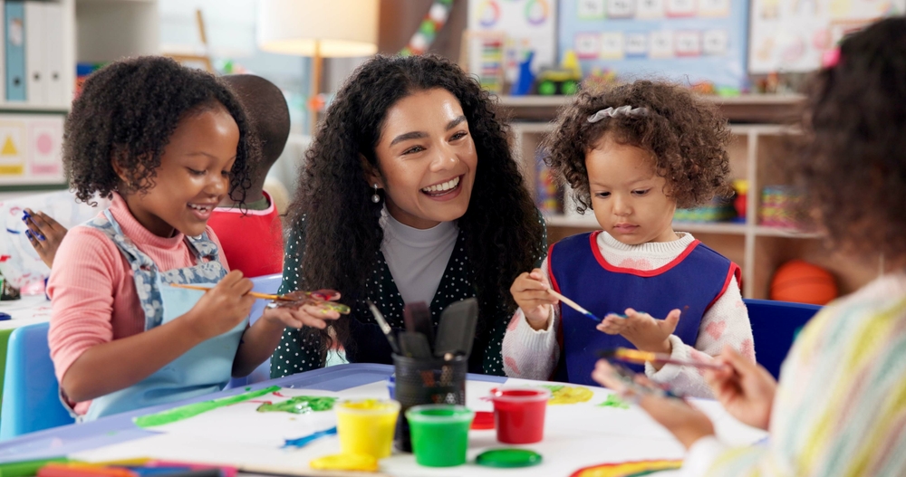EducationKidsAndTeacherPaintingHandsForArtCreativeTalent Preschool teacher guiding young children in a classroom art activity, with kids painting and exploring colors using handprints.