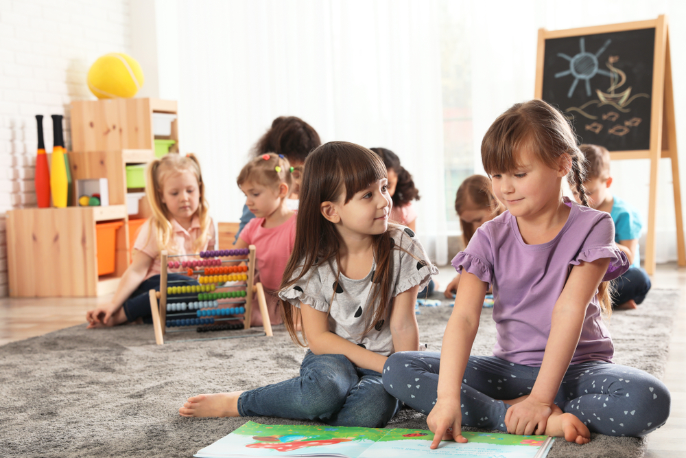 CuteGirlsReadingBookOnFloorWhileOtherChildrenPlaying Two preschool-aged girls sitting on the floor reading a book together and sharing a smile.