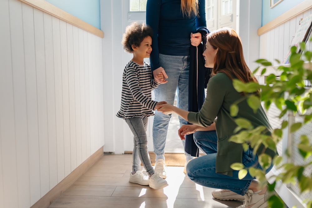PreschoolTeacherWarmlyWelcomesAChildAndHerMotherAt Preschool teacher warmly welcoming a child with her mother at school