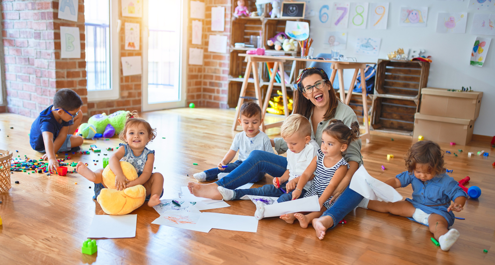 BeautifulTeacherAndGroupOfToddlersSittingOnTheFloor Preschool teacher sitting with a group of toddlers in a childcare classroom.